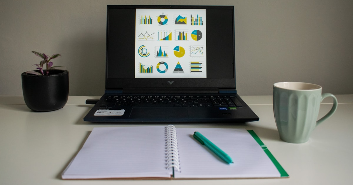 Laptop displaying charts beside a notebook and mug
